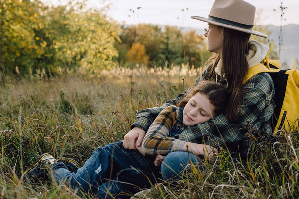Calm young mother and adorable little daughter lying in grass on background epic autumn mountains, enjoying beautiful nature during sunset. Family trip on fall vacation. Holiday and travel concept - Foto, Bild