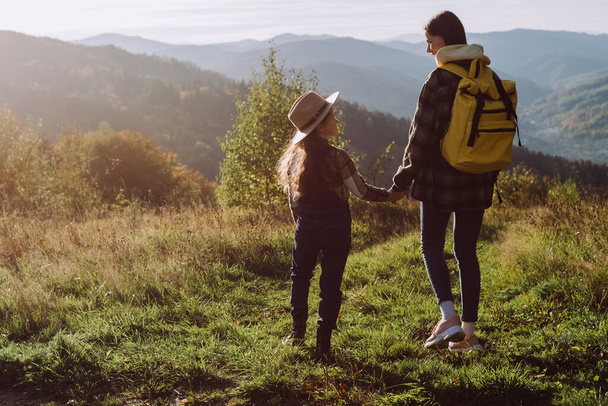 Traveler attractive young mother and pretty little daughter standing in grass on background beautiful calm autumn mountains at sunny day, enjoying nature. Lifestyle family travel tourism concept - Foto, Imagem