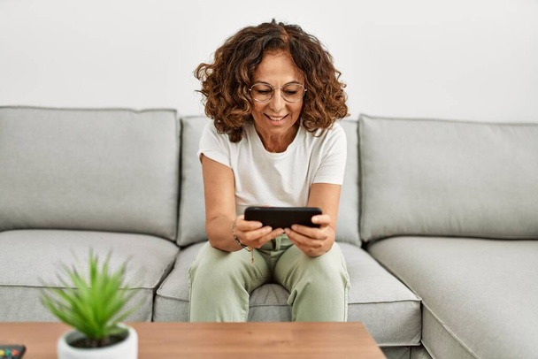 Mujer hispana de mediana edad sonriendo confiada usando smartphone en casa - Foto, imagen