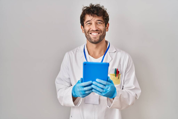 Hombre científico hispano trabajando con tableta que se ve positivo y feliz de pie y sonriendo con una sonrisa confiada mostrando los dientes  - Foto, Imagen