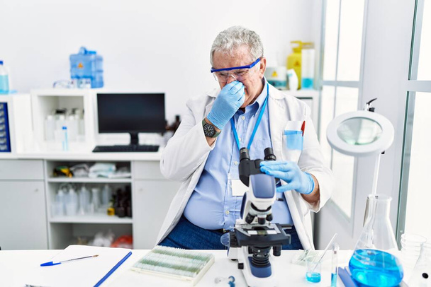 Starszy biały mężczyzna pracujący w laboratorium naukowym wyczuwa coś śmierdzącego i obrzydliwego, nieznośnego, wstrzymując oddech palcami na nosie. nieprzyjemny zapach  - Zdjęcie, obraz