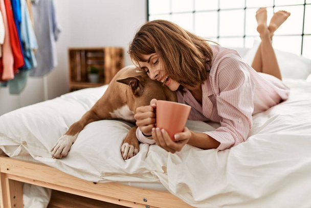 Young caucasian woman drinking coffee lying on bed with dog at bedroom - Фото, изображение