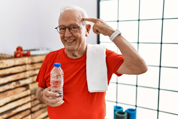 Starší muž ve sportovním oblečení a ručník v tělocvičně s úsměvem ukazuje na hlavu jedním prstem, skvělý nápad nebo myšlenka, dobrá paměť  - Fotografie, Obrázek