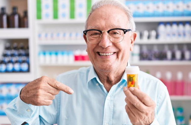 Hombre mayor con pelo gris sosteniendo pastillas en la farmacia señalando con el dedo a uno mismo sonriendo feliz y orgulloso  - Foto, imagen