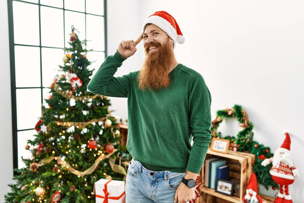 Homme rousse à longue barbe portant un chapeau de Noël par arbre de Noël souriant pointant vers la tête avec un doigt, grande idée ou pensée, bonne mémoire  - Photo, image