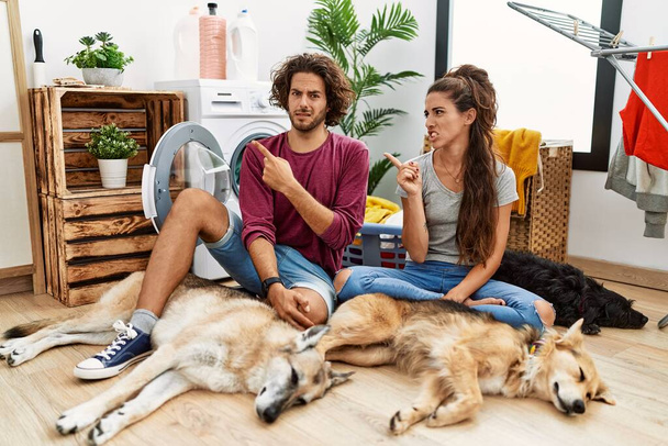 Young hispanic couple doing laundry with dogs pointing aside worried and nervous with forefinger, concerned and surprised expression  - Foto, Imagen
