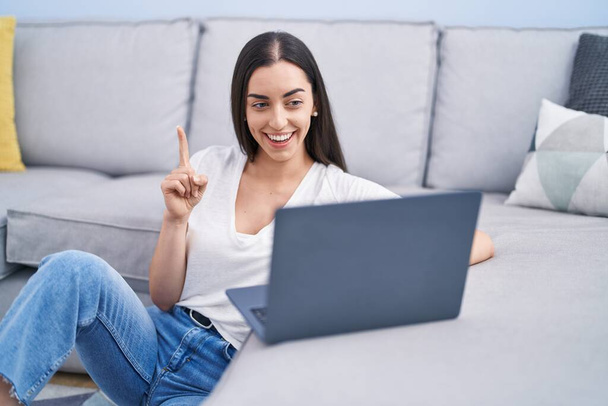 jonge brunette vrouw met behulp van laptop thuis glimlachen met een idee of vraag wijzend vinger met gelukkig gezicht, nummer een  - Foto, afbeelding