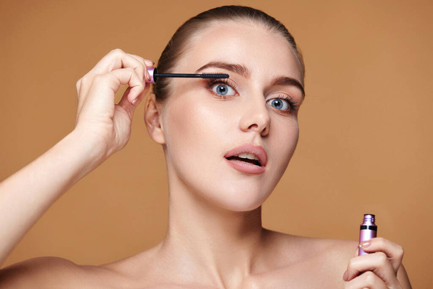 Beautiful woman applying black mascara on long thick eyelashes with brush and making makeup on beige background - Фото, изображение