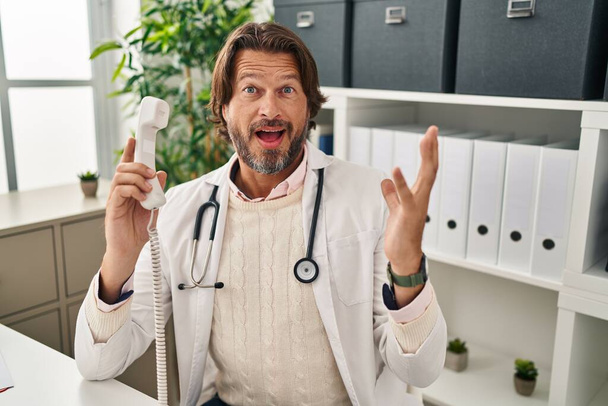 Handsome middle age doctor man speaking on the phone at the clinic celebrating victory with happy smile and winner expression with raised hands  - 写真・画像
