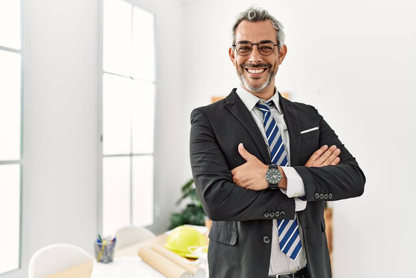 Middelbare leeftijd grijsharige man architect staat met armen gekruist gebaar op kantoor - Foto, afbeelding