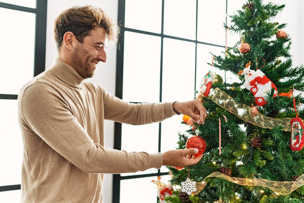 Joven hombre hispano sonriendo confiado decorando el árbol de Navidad en casa - Foto, imagen