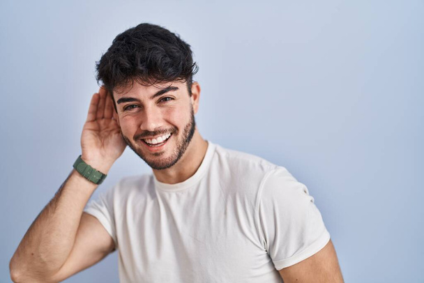 Latinomies, jolla on parta valkoisella taustalla hymyillen käsi korvassa kuunnellen huhuja tai juoruja. kuurous käsite.  - Valokuva, kuva