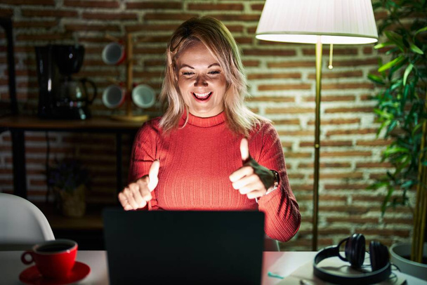 Mulher loira usando laptop à noite em casa aprovando fazer gesto positivo com a mão, polegares para cima sorrindo e feliz pelo sucesso. gesto vencedor.  - Foto, Imagem