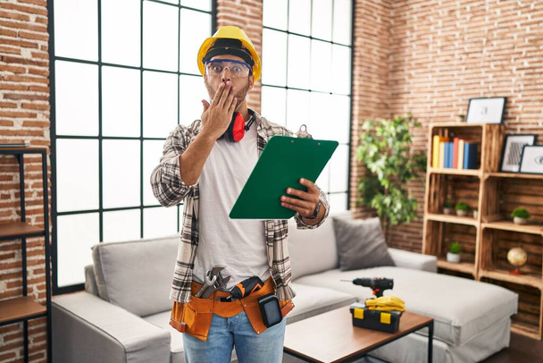 Young hispanic man with beard working at home renovation covering mouth with hand, shocked and afraid for mistake. surprised expression  - 写真・画像