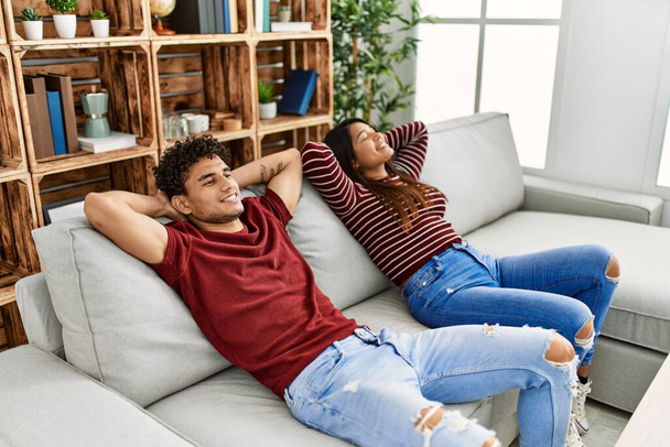 Νεαρό latin ζευγάρι χαμογελά ευτυχισμένη χαλαρή με τα χέρια στο κεφάλι κάθεται στον καναπέ στο σπίτι. - Φωτογραφία, εικόνα