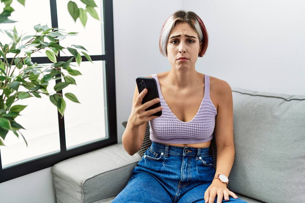 Junge schöne Frau mit Smartphone-Tippnachricht sitzt deprimiert auf dem Sofa und sorgt sich vor Kummer, weint wütend und ängstlich. Traurige Miene.  - Foto, Bild