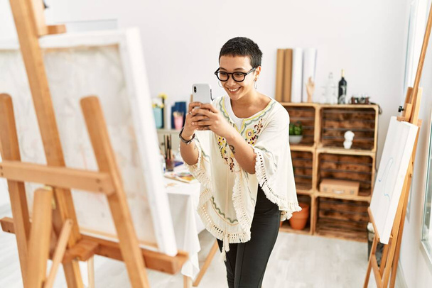 Mujer hispana joven hacer foto por el teléfono inteligente para dibujar lienzo en el estudio de arte - Foto, Imagen