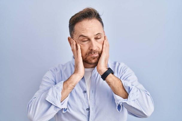 Middle age caucasian man standing over blue background tired hands covering face, depression and sadness, upset and irritated for problem  - Foto, Bild