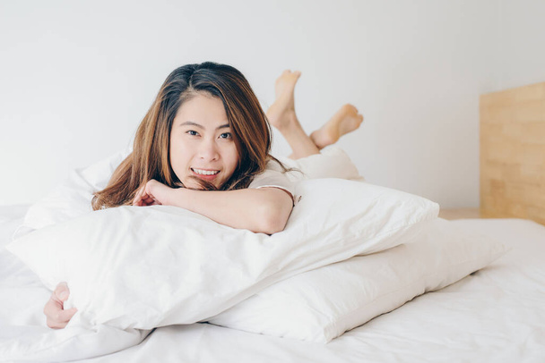 Szczęśliwa Azjatka leżąca na łóżku ze skrzyżowanymi nogami i patrząca do kamery. - Zdjęcie, obraz