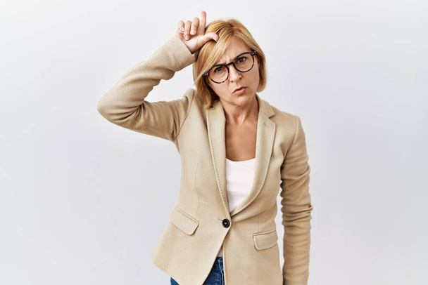 Middle age blonde business woman standing over isolated background making fun of people with fingers on forehead doing loser gesture mocking and insulting.  - Foto, imagen