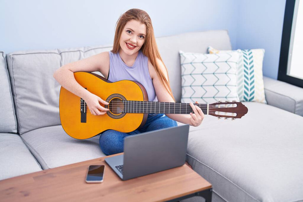Genç kızıl saçlı kadın evdeki kanepede oturmuş online klasik gitar dersi alıyor. - Fotoğraf, Görsel