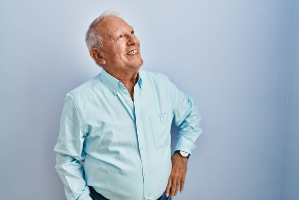 Hombre mayor con el pelo gris de pie sobre fondo azul mirando hacia un lado con sonrisa en la cara, expresión natural. riendo confiado.  - Foto, imagen