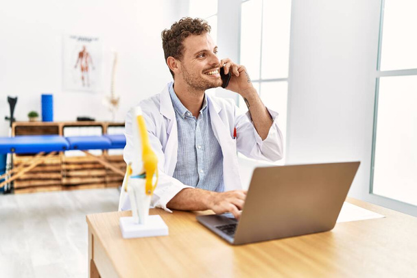Giovane uomo ispanico indossa uniforme fisioterapista utilizzando computer portatile parlando sullo smartphone in clinica - Foto, immagini