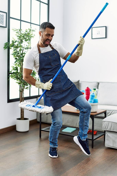 Young hispanic man cleaning floor and singing song using mop as a guitar at home - Foto, Imagen