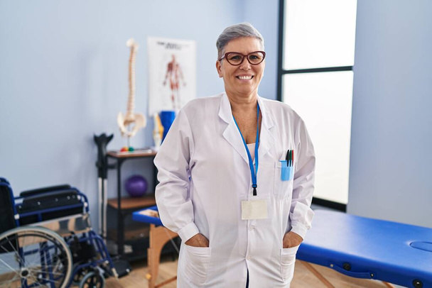 Fisioterapista donna di mezza età sorridente fiducioso in piedi alla clinica di riabilitazione - Foto, immagini