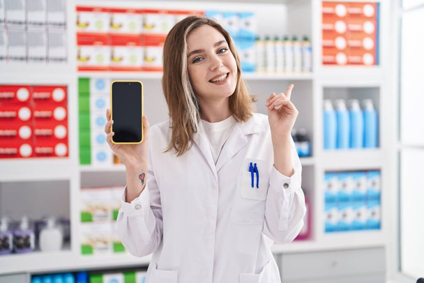 Blonde caucasian woman working at pharmacy drugstore showing smartphone screen smiling happy pointing with hand and finger to the side  - Foto, Imagem