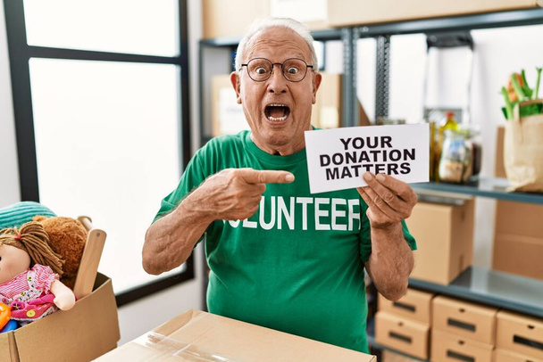Hombre voluntario mayor sosteniendo su donación importa sonriendo feliz señalando con la mano y el dedo  - Foto, imagen