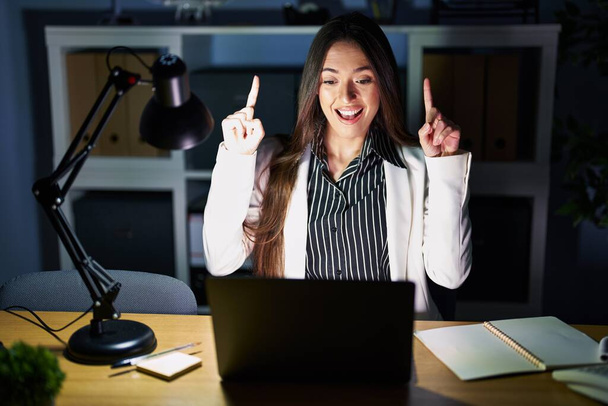Młoda brunetka pracująca w biurze w nocy z laptopem uśmiechnięta, zaskoczona i wskazująca palcami i podniesionymi ramionami.  - Zdjęcie, obraz