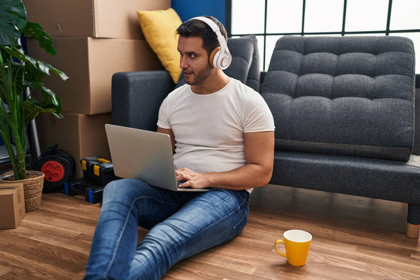 Молодой латиноамериканец использует ноутбук, сидя на полу в новом доме - Фото, изображение
