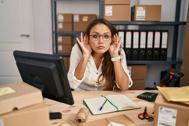 Young hispanic woman working at small business ecommerce trying to hear both hands on ear gesture, curious for gossip. hearing problem, deaf  - 写真・画像