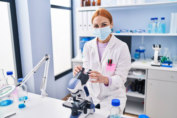 Fiatal fehér tudós nő orvosi maszkot visel mikroszkóppal a laboratóriumban - Fotó, kép