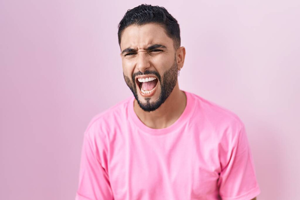 Ισπανόφωνος νεαρός άνδρας στέκεται πάνω από ροζ φόντο θυμωμένος και τρελός ουρλιάζοντας απογοητευμένος και έξαλλος, φωνάζοντας από θυμό. οργή και επιθετική αντίληψη.  - Φωτογραφία, εικόνα