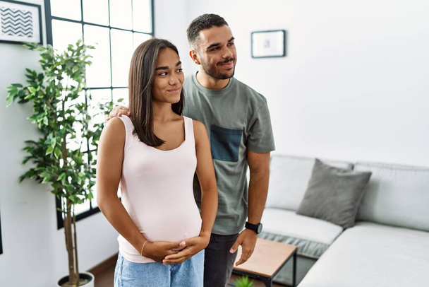 Jovem casal interracial esperando um bebê, tocando barriga grávida sorrindo olhando para o lado e olhando para longe pensando.  - Foto, Imagem