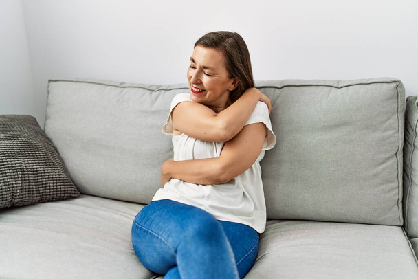Middle age hispanic woman smiling sitting on the sofa hugging herself at home - Photo, Image
