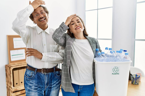 Mittelalterliches gemischtes Paar, das im Büro gestresst und frustriert Recyclingbehälter mit Plastikflaschen in der Hand hält, überrascht und wütend  - Foto, Bild