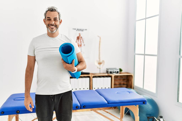 Middle age grey-haired man rehab patient holding yoga mat at rehab clinic - Foto, Imagem