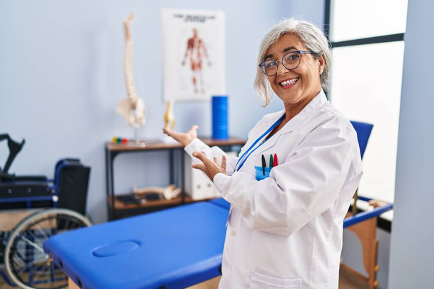 Donna di mezza età con i capelli grigi che lavora alla clinica di recupero del dolore invitando ad entrare sorridendo naturale a mano aperta  - Foto, immagini