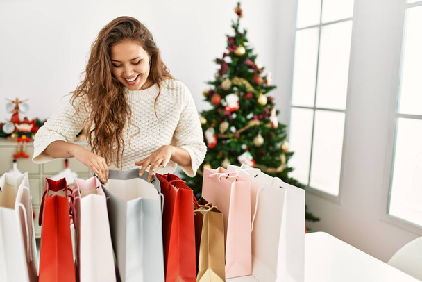 Nuori kaunis latino nainen etsii ostoskassit seisoo joulukuusi kotona - Valokuva, kuva