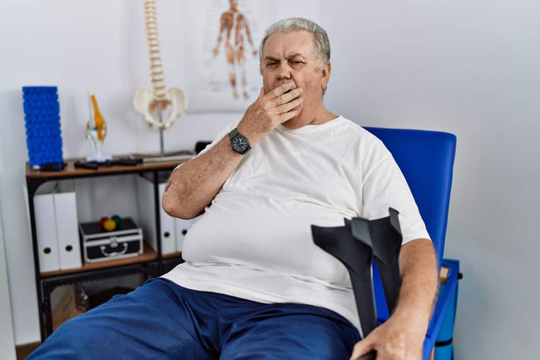 Starszy biały mężczyzna w klinice fizjoterapeutycznej trzymający kule znudzony ziewaniem zmęczonym zakrywaniem ust ręką. niepokój i senność.  - Zdjęcie, obraz