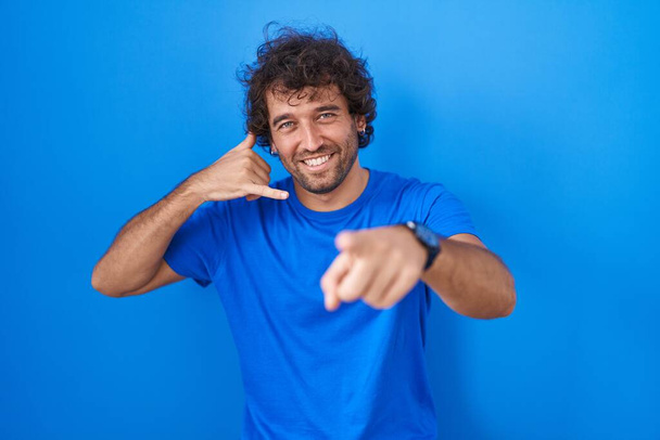 Hispanic young man standing over blue background smiling doing talking on the telephone gesture and pointing to you. call me.  - Photo, Image