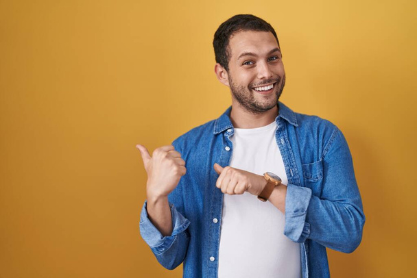 Hispanic man standing over yellow background pointing to the back behind with hand and thumbs up, smiling confident  - Photo, image