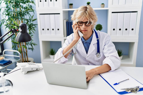 młody blond mężczyzna lekarz za pomocą laptopa rozmowy przez telefon w klinice - Zdjęcie, obraz