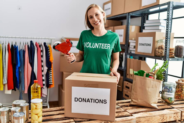 Jeune femme caucasienne portant bénévole uniforme emballage boîte en carton travaillant au centre de charité - Photo, image