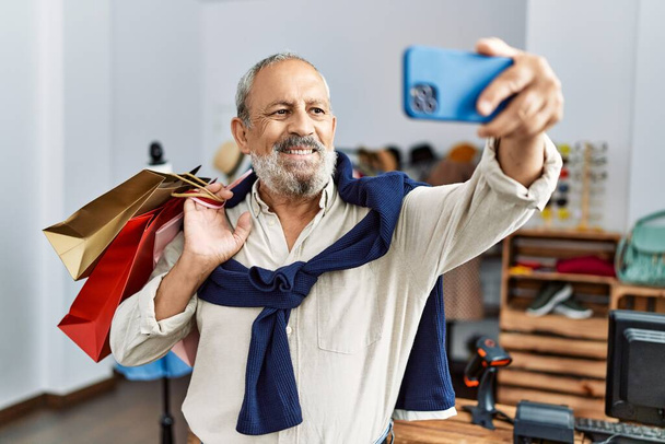 Starší šedovlasý muž držící nákupní tašky dělá selfie u smartphonu v obchodě s oblečením - Fotografie, Obrázek
