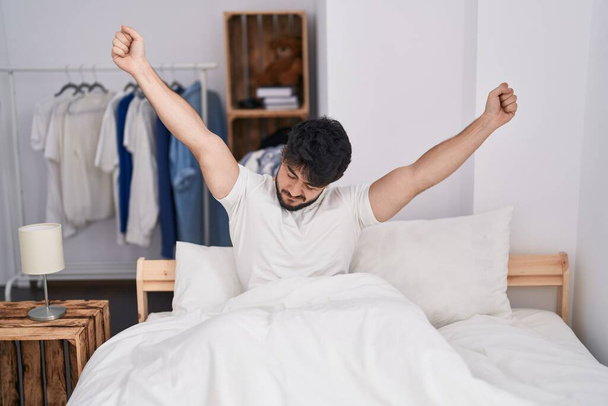 Young hispanic man waking up stretching arms at bedroom - Foto, imagen