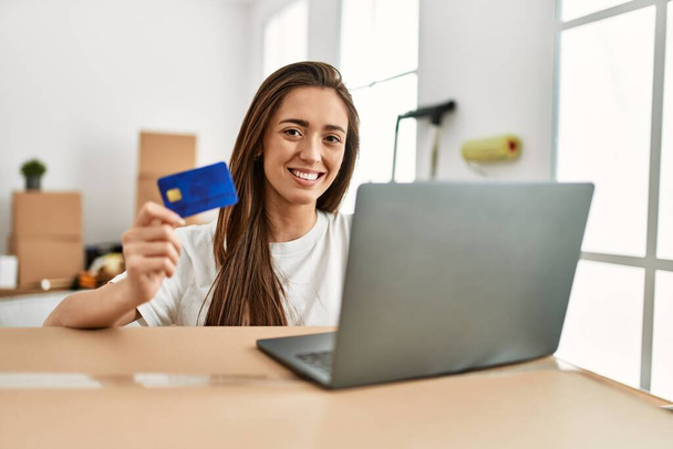 Молода іспаномовна жінка посміхається впевнено використовуючи кредитну картку і ноутбук в новому будинку
 - Фото, зображення